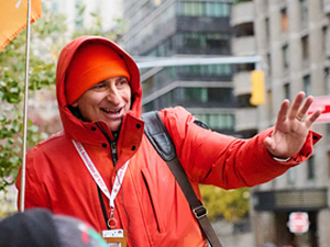 Jared leading a group in his signature orange jacket, holding a flag on a Manhattan street